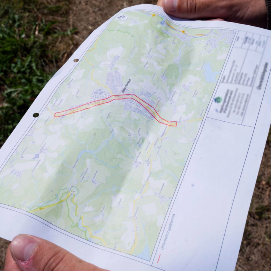 Zwei Hände halten einen Übersichtslagenplan mit rot markiertem Trassenverlauf durch Breckerfeld, im Hintergrund Gras und Erdboden der Baustelle.